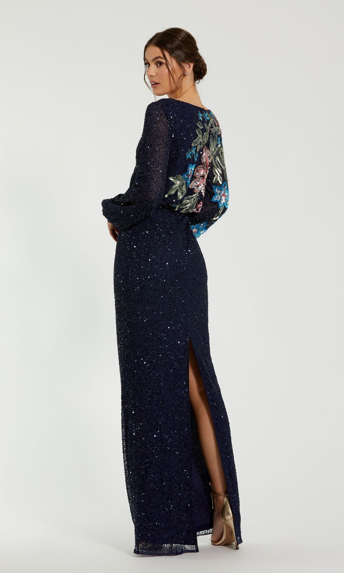 A woman stands with her back turned and head over her shoulder, wearing the Mac Duggal 5615 Beaded Long Sleeve Formal Dress, showcasing navy blue sequin details and floral embroidery on the back.